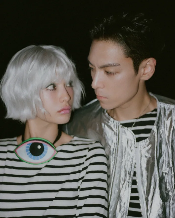An artistic fashion photograph of Nana with short silver hair wearing a striped top and a vibrant eye-shaped necklace, standing close to T.O.P with dark hair in a shiny silver jacket, both gazing at each other against a dark background.