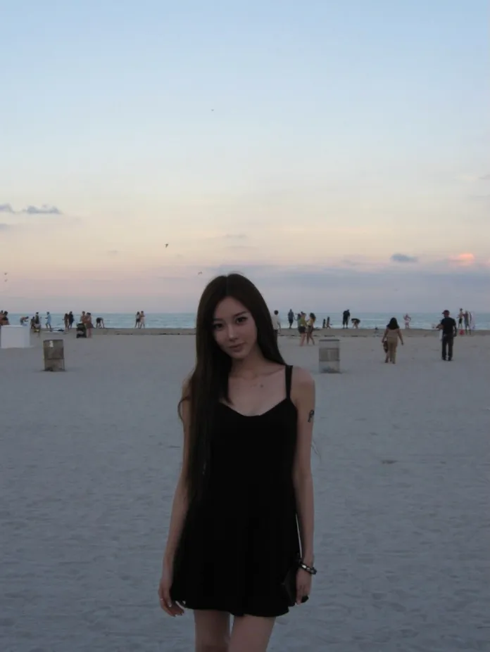 Giselle  in a black dress standing on a beach at sunset, with people in the background and a pastel sky.