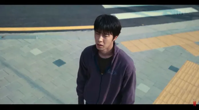 Lee Jae-wook standing on a crosswalk, looking upwards with a concerned expression. He is wearing a purple jacket and the ground is paved with light-colored tiles and yellow tactile paving.