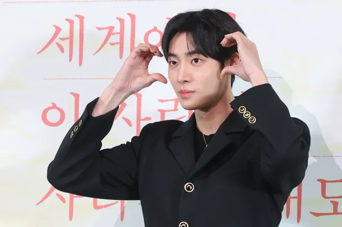 Choo Young-woo in a black suit, making a playful gesture with his hands while posing for the camera.