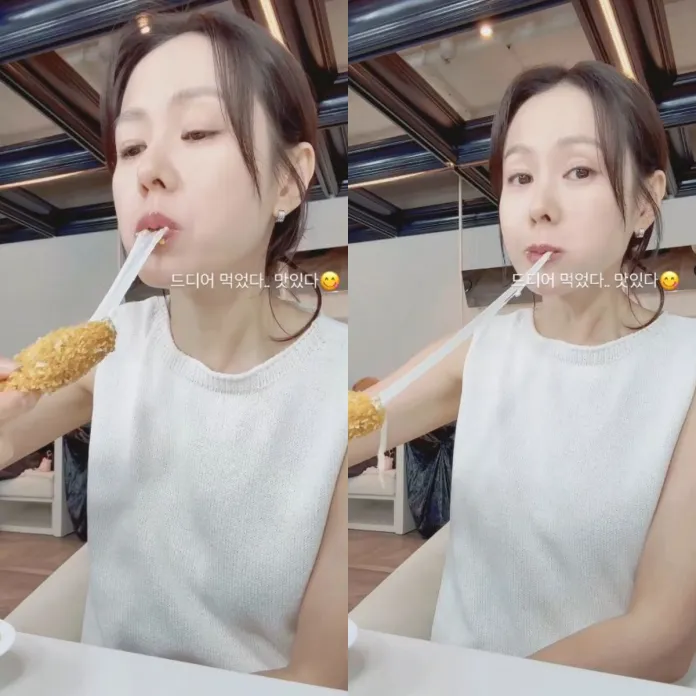 Son Ye-jin enjoying a food item on a stick with chopsticks, looking pleased while seated in a casual dining setting.