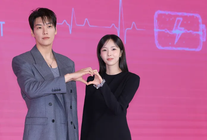 Two individuals standing together making a heart shape with their hands, with a pink background featuring a heartbeat line and battery icon.