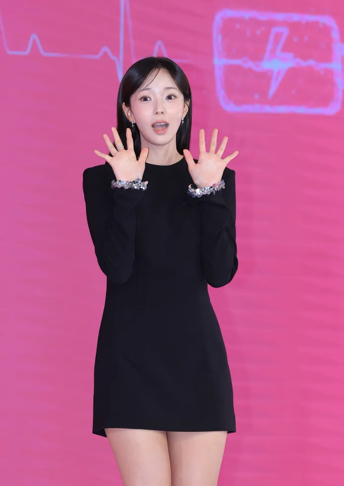 A woman in a black dress is performing with her hands raised in front of a pink background featuring a heartbeat graphic.