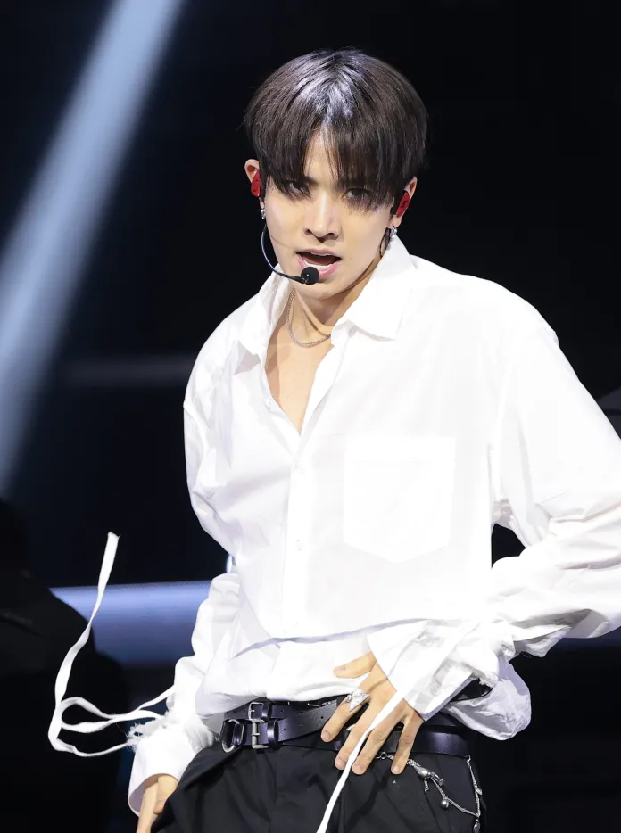 A young performer in a white shirt with rolled-up sleeves, wearing a microphone headset, poses confidently on stage with dramatic lighting.