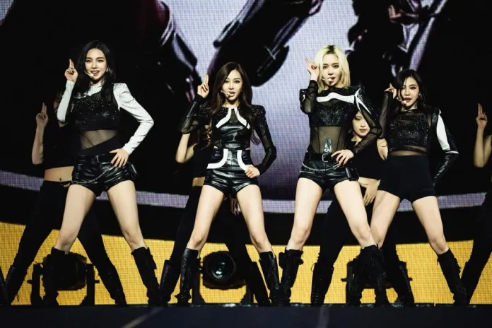 Aespa members performing on stage in stylish outfits, featuring black shorts and sheer tops, showcasing synchronized dance moves.