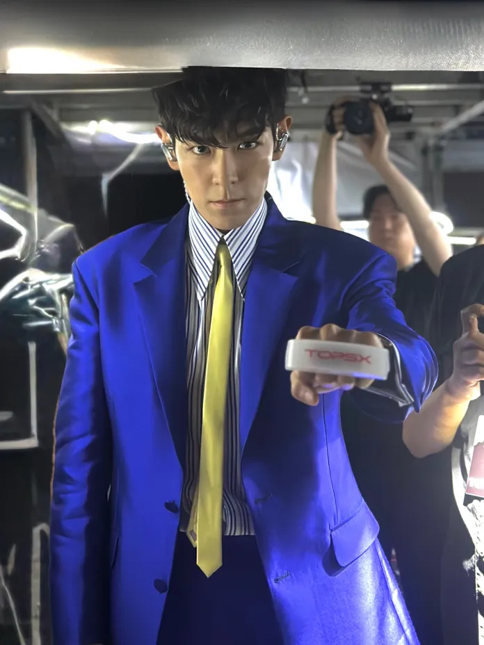 T.O.P in a striking blue suit with a yellow tie, holding a device labeled 'TOPSX' and looking intently at the camera. The background features blurred figures, indicating a lively environment.
