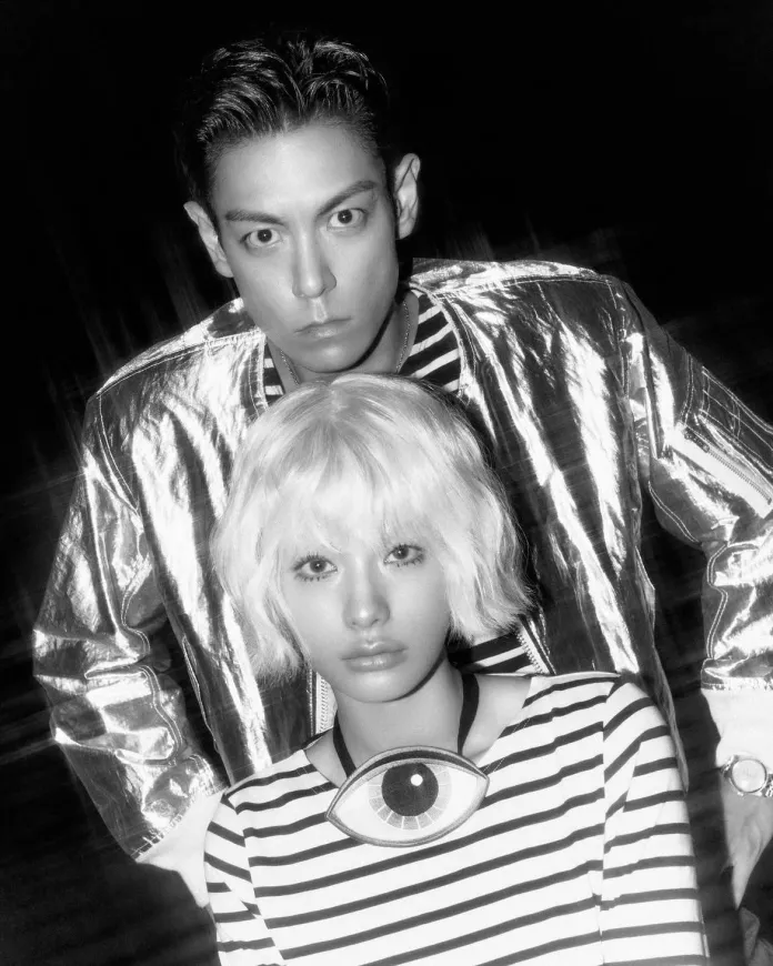 A black and white photo featuring a T.O.P in a shiny silver jacket and a NANA with short, wavy blonde hair wearing a striped shirt with an eye-shaped pendant.