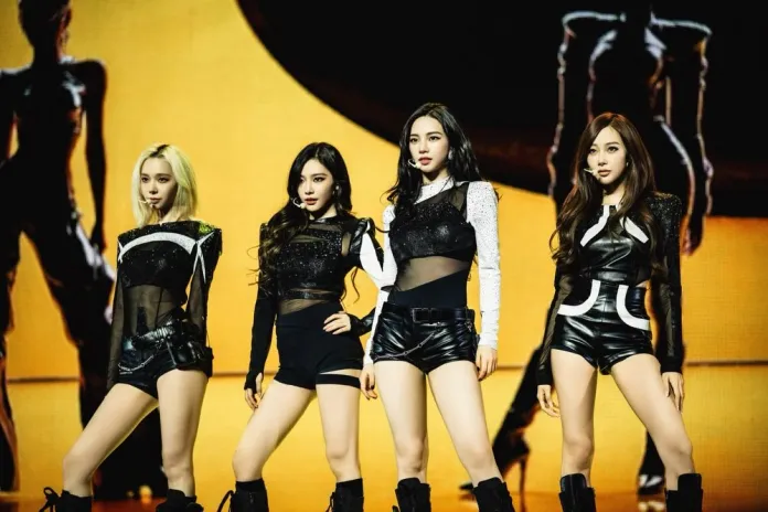 aespa members standing confidently on stage, wearing stylish black outfits and posing strikingly. The background features dramatic lighting and silhouettes.