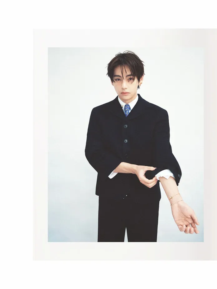 BTS's V in a fitted black suit rolls up his shirt sleeves while looking at the camera with a serious expression.