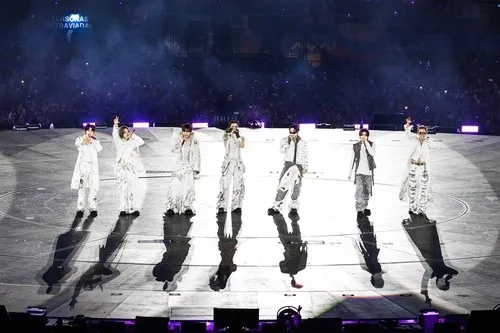 BTS on stage wearing white outfits, waving to the audience under bright lights during a concert.