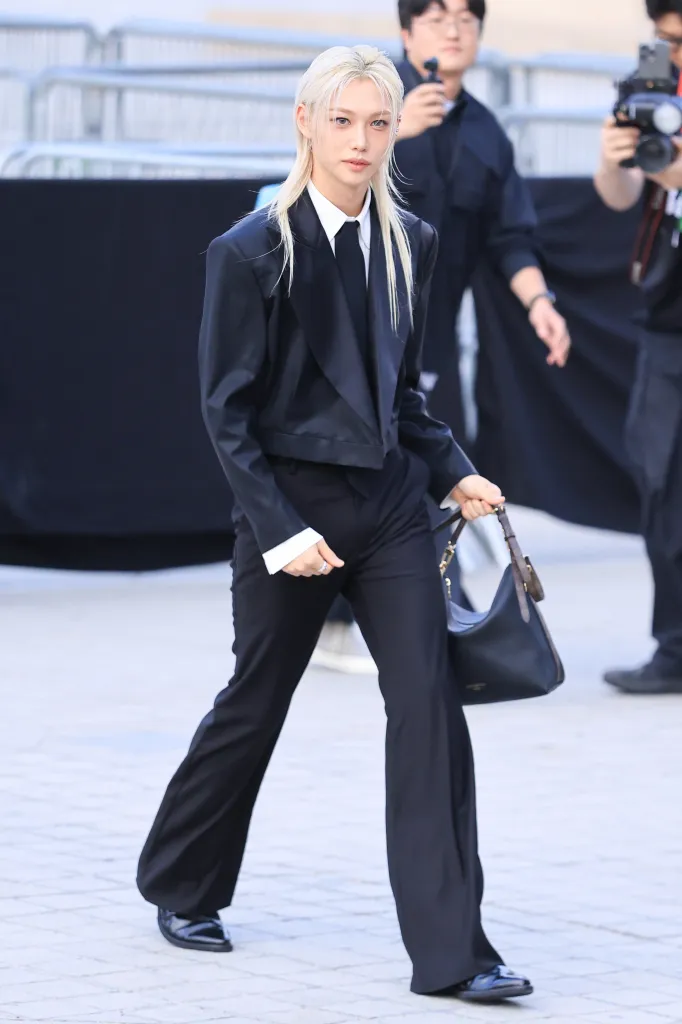 Stray Kids’ Felix with long blonde hair wearing a black suit, white shirt, and tie, walking confidently while carrying a handbag. Photographers can be seen in the background.