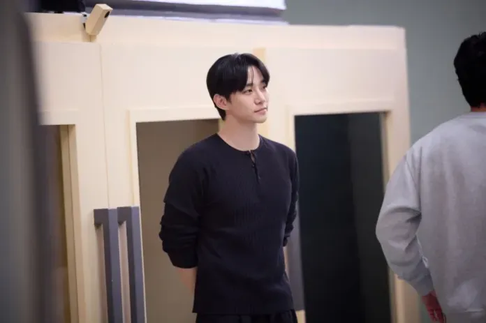 Lee Junho with dark hair stands casually in a room, wearing a black long-sleeve shirt. He appears contemplative, looking to the side, while another person in a gray outfit is partially visible in the background.