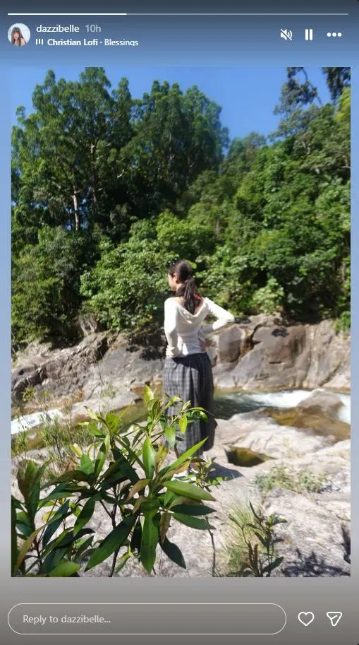 Hanni standing on a rocky riverbank, facing away with hands on hips, surrounded by lush greenery and flowing water.