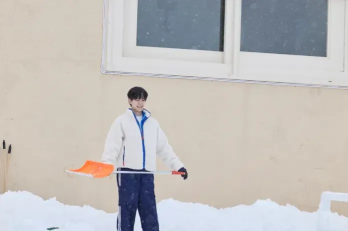 Lee Junho standing in the snow holding an orange shovel, wearing winter clothing, with a house in the background and snow falling.