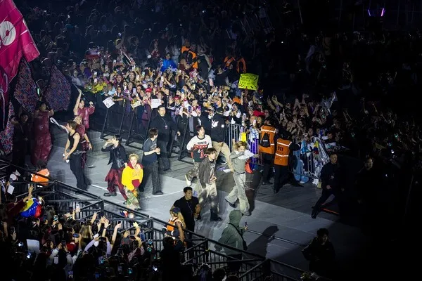 BTS with performers on stage engaging with fans. The audience is holding signs and cheering, while security personnel are visible in the foreground. Bright stage lighting enhances the lively atmosphere.