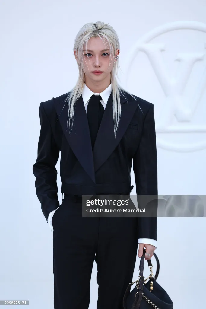 Stray Kids’ Felix wearing a stylish black suit with a white shirt and tie, standing confidently while holding a designer handbag.
