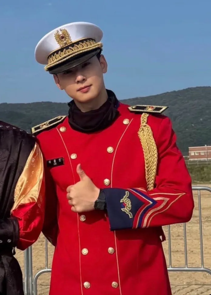 Cha Eun Woo in a red military-style uniform with gold accents and a white peaked cap gives a thumbs-up gesture. He is standing outdoors with a mountainous background.