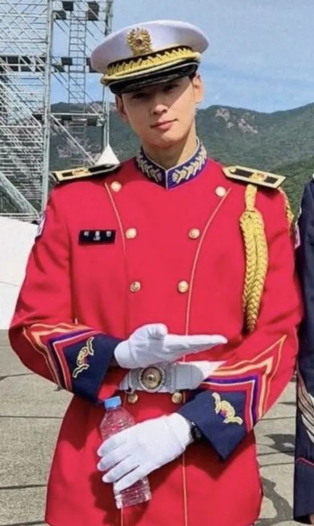 Cha Eun Woo in a formal red military uniform with gold accents and a white hat, holding a water bottle and posing with one hand in front of him.