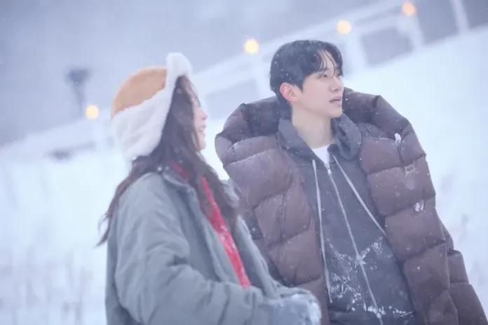 Lee Junho in a winter coat and hat and a young man in a puffy jacket stand together in a snowy landscape, with snow gently falling around them.