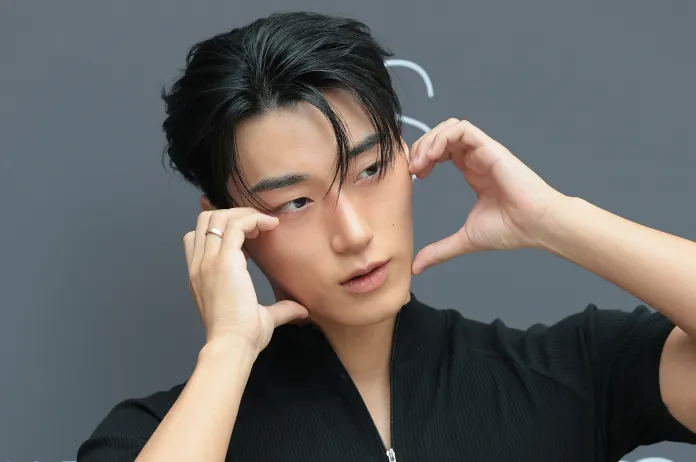 ATEEZ's San in a black polo shirt poses with fingers touching his face, expressing contemplation against a gray background.