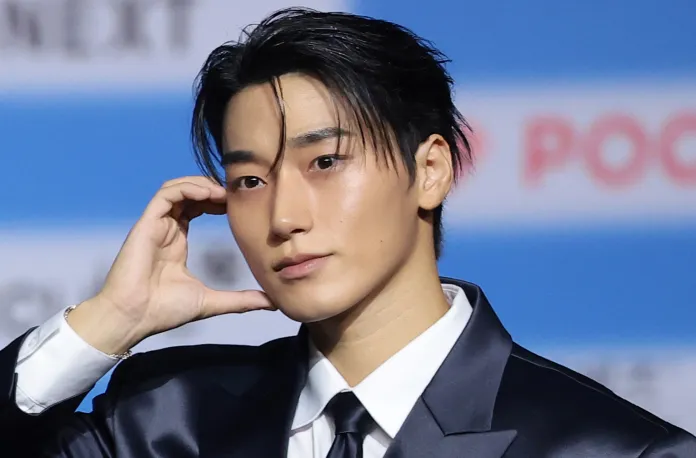 ATEEZ's San in a formal black suit poses confidently with one hand near his face, showcasing a stylish hairstyle and a thoughtful expression.