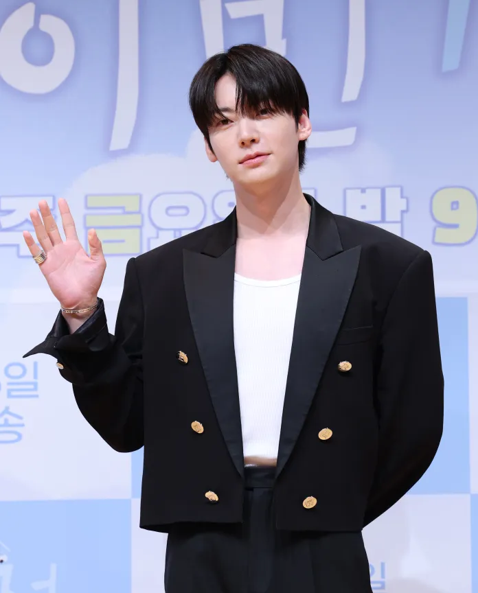 A stylish individual waves at the camera, wearing a black blazer with gold buttons over a white shirt, against a light blue background with text.