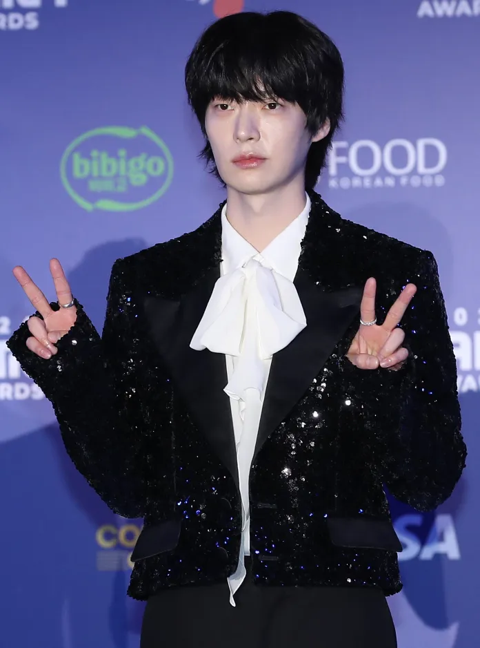 A young man wearing a sequined black jacket and a white bow tie poses on the red carpet, making a peace sign with both hands.