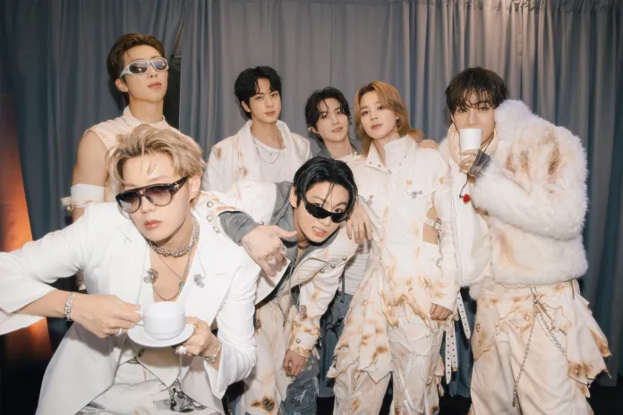 BTS posing together in a stylish setting. They are dressed in coordinated fashion outfits featuring light colors and unique textures. The group exhibits a mix of casual and high-fashion styles, with some members wearing sunglasses and holding cups.
