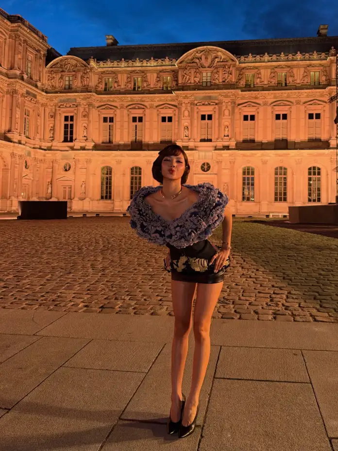 A woman stands in front of a grand historical building at dusk, wearing a stylish outfit with a floral design. She is posing playfully with a pout and has a confident stance, surrounded by a cobblestone area.