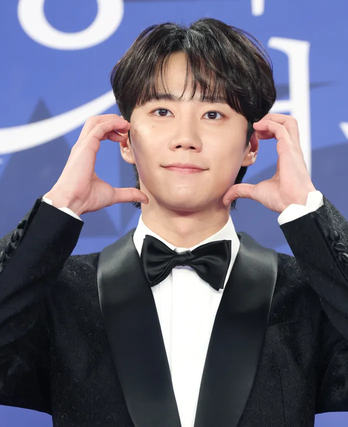 Lee Jun-young in a tuxedo with a bow tie making a playful gesture by touching his face, posing at a formal event with a blue backdrop.