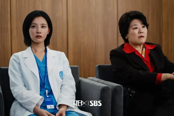 Kim Ji-won in a white coat sitting next to Lee Jung-eun in a red and black outfit, both appearing serious in a modern office setting.