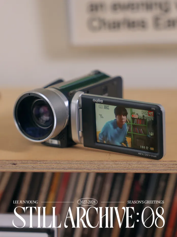 A camcorder placed on a wooden surface, with its screen displaying Lee Jun-young in a blue sweater. In the background, there are shelves with books or vinyl records. The text overlay reads 'LEE JUN YOUNG 2027-2028 SEASON'S GREETINGS STILL ARCHIVE: 08'.