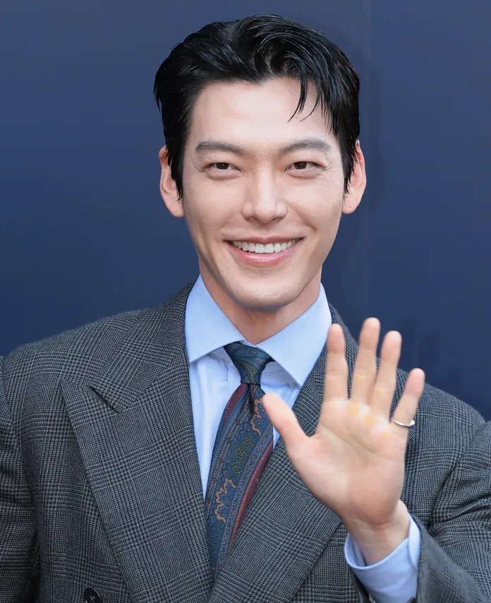 Kim Woo-bin in a stylish suit waves at the camera, with a neutral background.