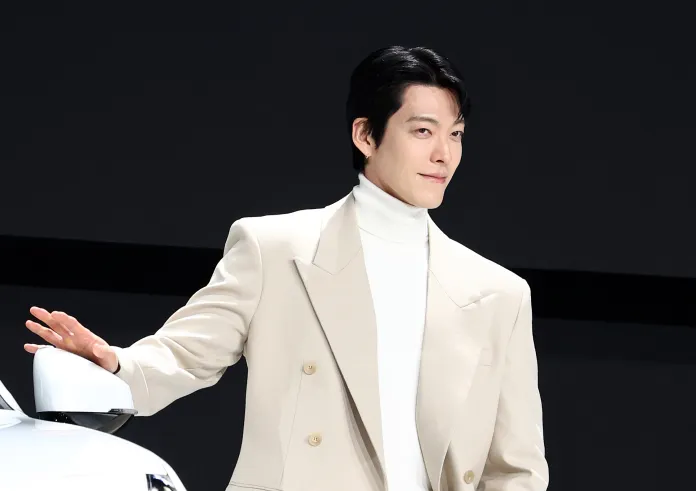 Kim Woo-bin in a beige suit and white turtleneck stands next to a white car, smiling and gesturing with their hand.