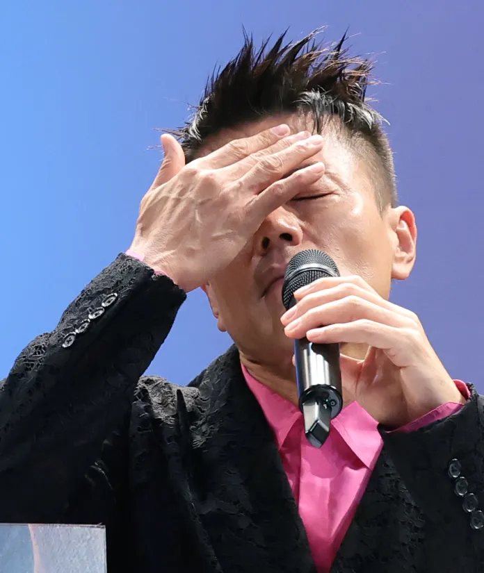 A man wearing a black patterned jacket and pink shirt is holding a microphone and partially covering his face with one hand, appearing to be in a moment of emotional expression.