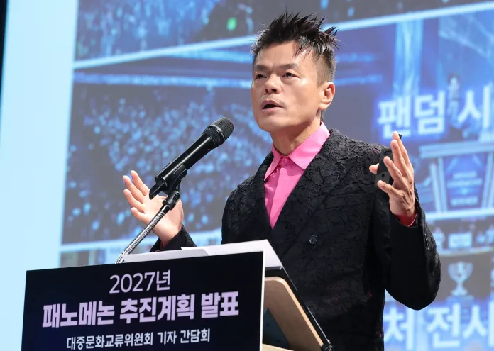 A man speaking at a podium during a presentation, wearing a black jacket and a pink shirt, with a focused expression and raised hand.