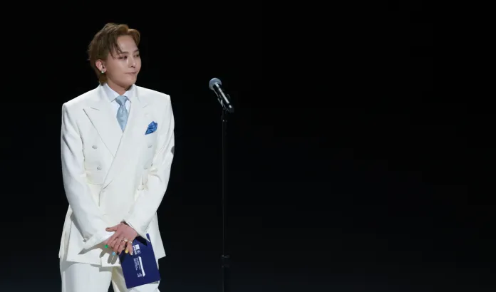 G-Dragon in a white suit with a light blue tie stands on stage next to a microphone, looking slightly to the side with a calm expression.