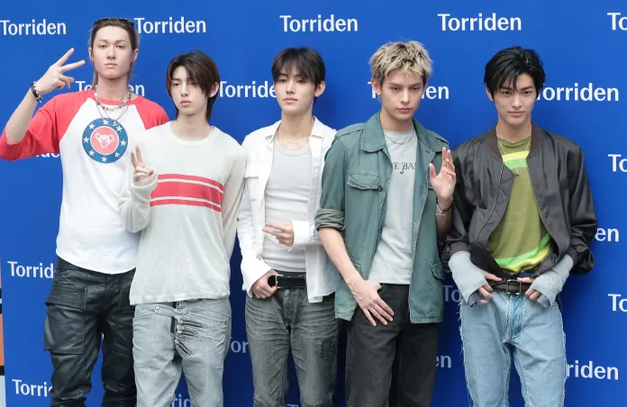 CORTIS members posing together in front of a blue backdrop with the word 'Torriden' printed. They display casual fashion styles, with some making hand gestures.