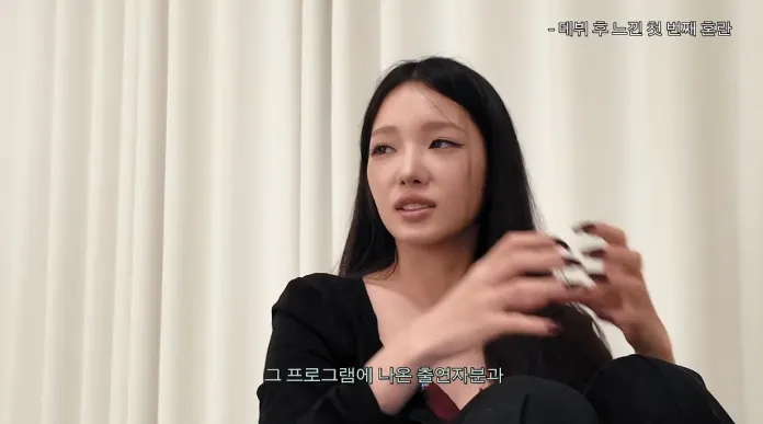 Roh Jisun sitting in front of a white curtain, holding a small object in her hands and engaging in conversation.