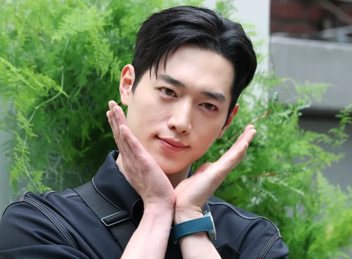 Seo Kang-joon with dark hair poses playfully with his hands under his chin, smiling softly, set against a backdrop of lush green foliage.