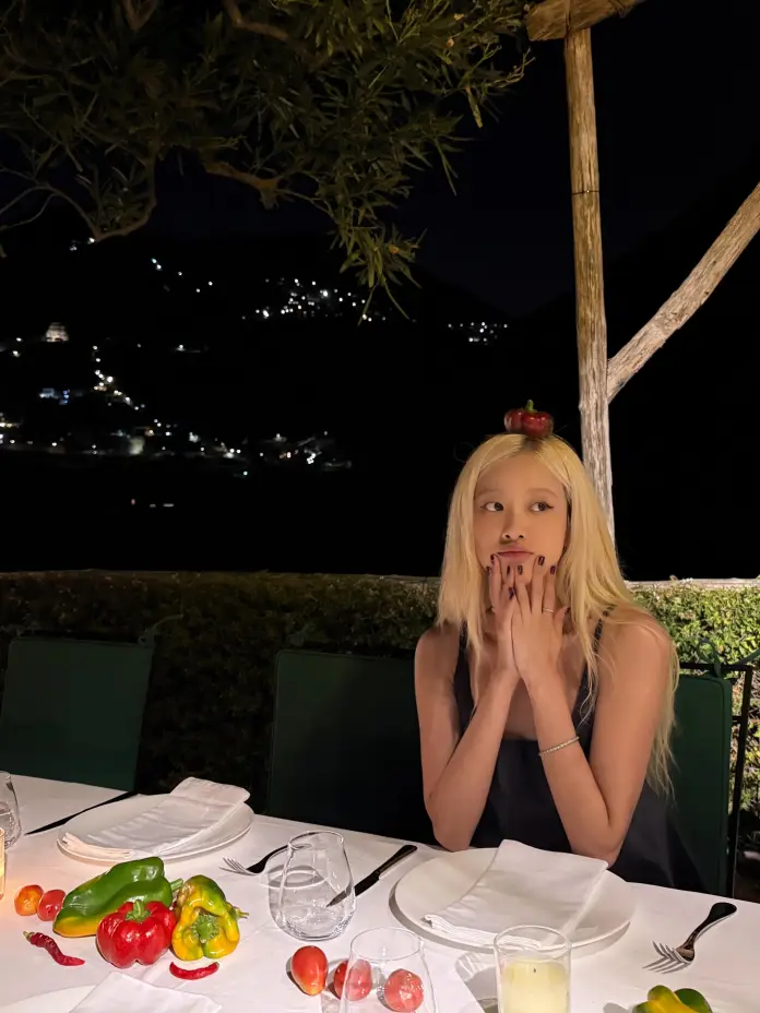 Rose sitting at a beautifully set dinner table outdoors at night, surrounded by colorful peppers and tomatoes, with a small red pepper on her head.