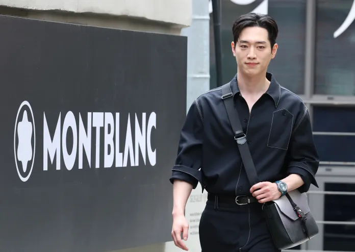 Seo Kang-joon walking in front of a Montblanc sign, wearing a black outfit and a watch, carrying a shoulder bag.