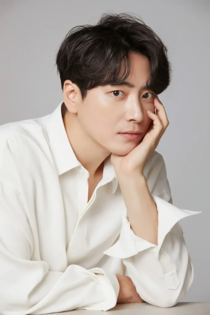 Lee Jun-hyuk with short, wavy black hair is seated, resting their chin on their hand. They are wearing a white shirt and looking directly at the camera with a neutral expression.