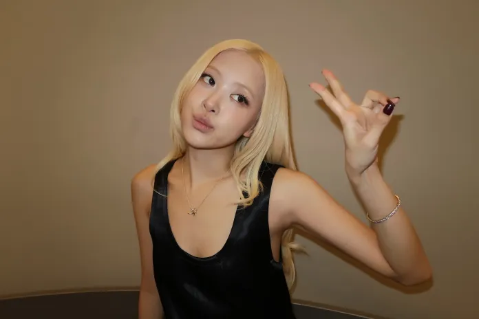 Rosé with long blonde hair wearing a black sleeveless top, posing with a playful hand gesture against a neutral background.