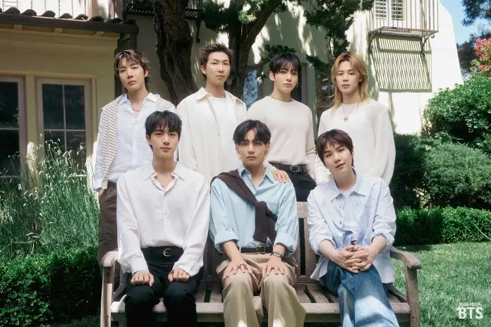 A group portrait of BTS members sitting outdoors on a wooden bench in a garden setting, featuring a mix of styles and casual attire.