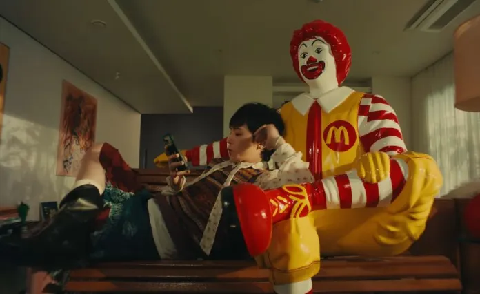 G-Dragon lounging on a couch with their phone, sitting next to a large Ronald McDonald statue.