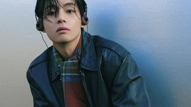 BTS V wearing a leather jacket and headphones, looking thoughtfully at the camera in a softly lit environment.