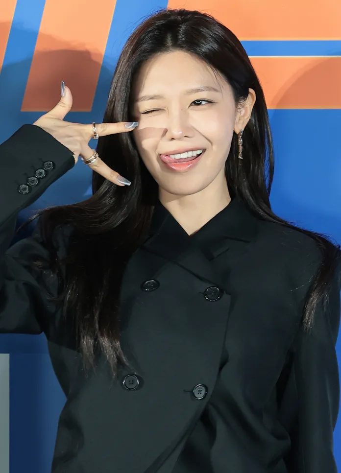 Choi Soo-young in a black double-breasted coat winks and makes a playful gesture with her hand, sticking out her tongue against a colorful background.
