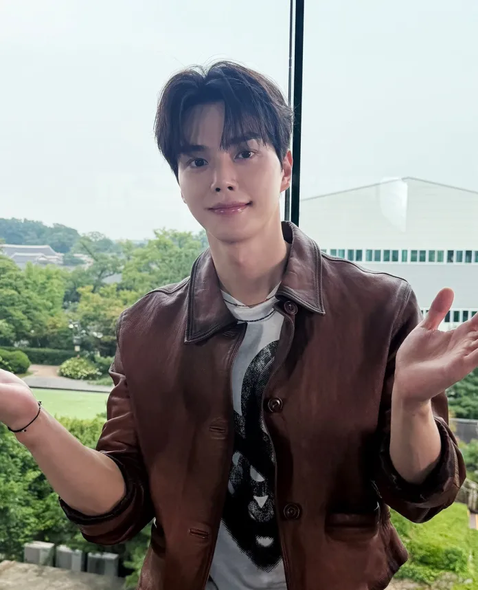 Song Kang wearing a brown leather jacket and a graphic t-shirt, smiling and gesturing with both hands while standing in front of a window with greenery visible outside.