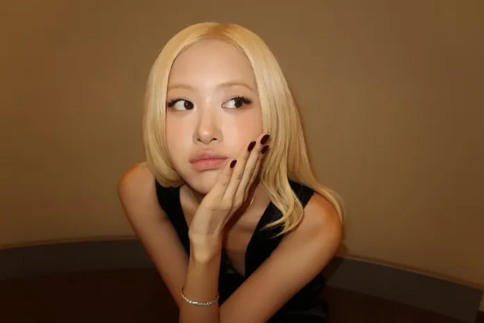 Rosé with long blonde hair is resting her chin on her hand, looking thoughtfully to the side. She is wearing a black outfit and has stylish, dark nail polish.